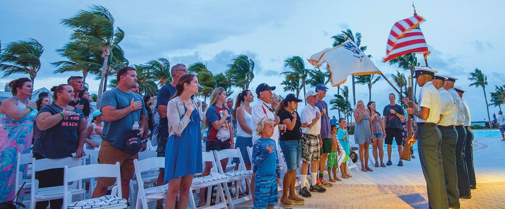 A group of people stands with their hands over their hearts during a flag ceremony near a pool with palm trees swaying in the background.