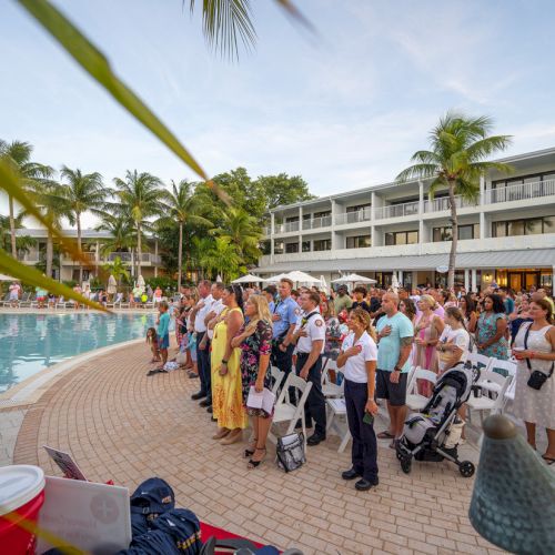 A group of people is gathered for an outdoor event by a pool, surrounded by palm trees and buildings, seemingly at a resort during the day.