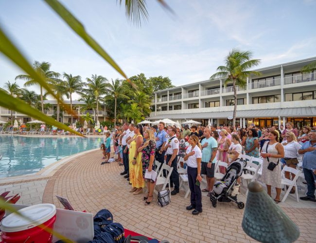 A group of people is gathered for an outdoor event by a pool, surrounded by palm trees and buildings, seemingly at a resort during the day.