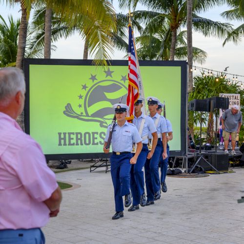 A group of uniformed individuals carrying the American flag marches past a screen displaying the word 