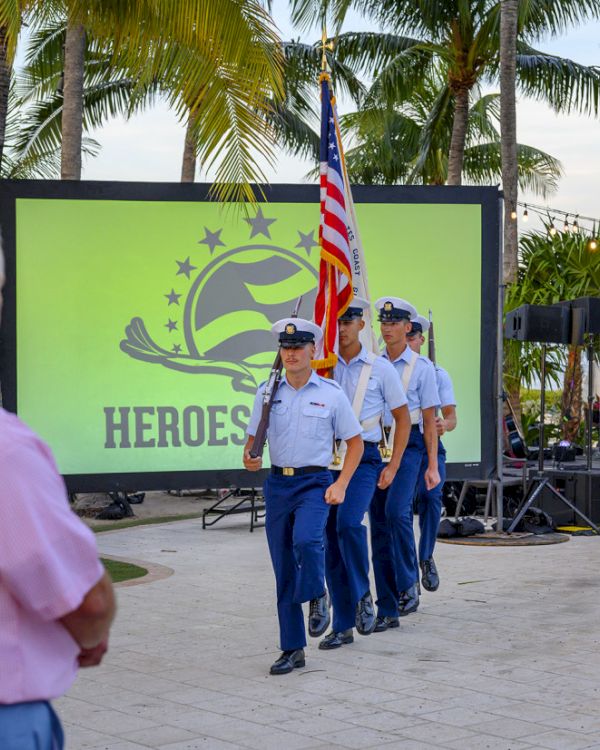 A group of uniformed individuals carrying the American flag marches past a screen displaying the word 